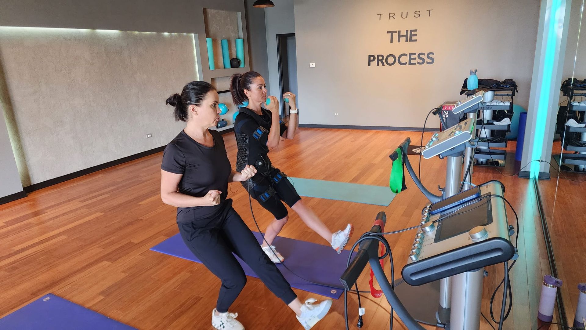 Two people using rowing machines in a studio with wooden floors and a wall sign reading “THE PROCESS.”