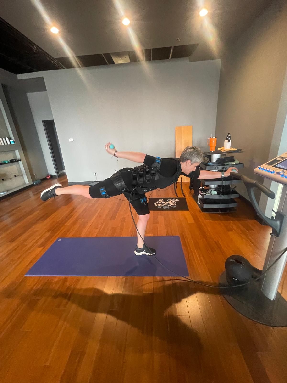 Person balancing on one leg on a purple mat in a wood-floored room with workout equipment.