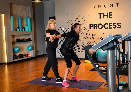 Two people exercising with resistance bands beside treadmills in a gym, with TRUST THE PROCESS on the wall.