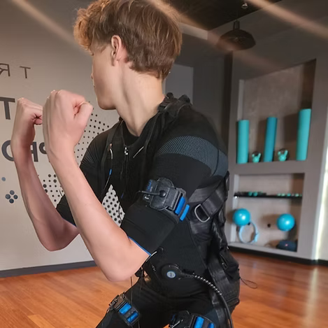 Person in a black EMS training suit flexing arms in a gym studio with wood floor and blue exercise balls