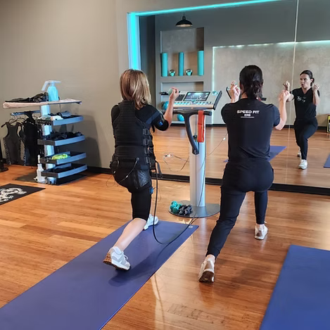 Two people exercising on stationary bikes in a mirrored studio with wood floors and yoga mats