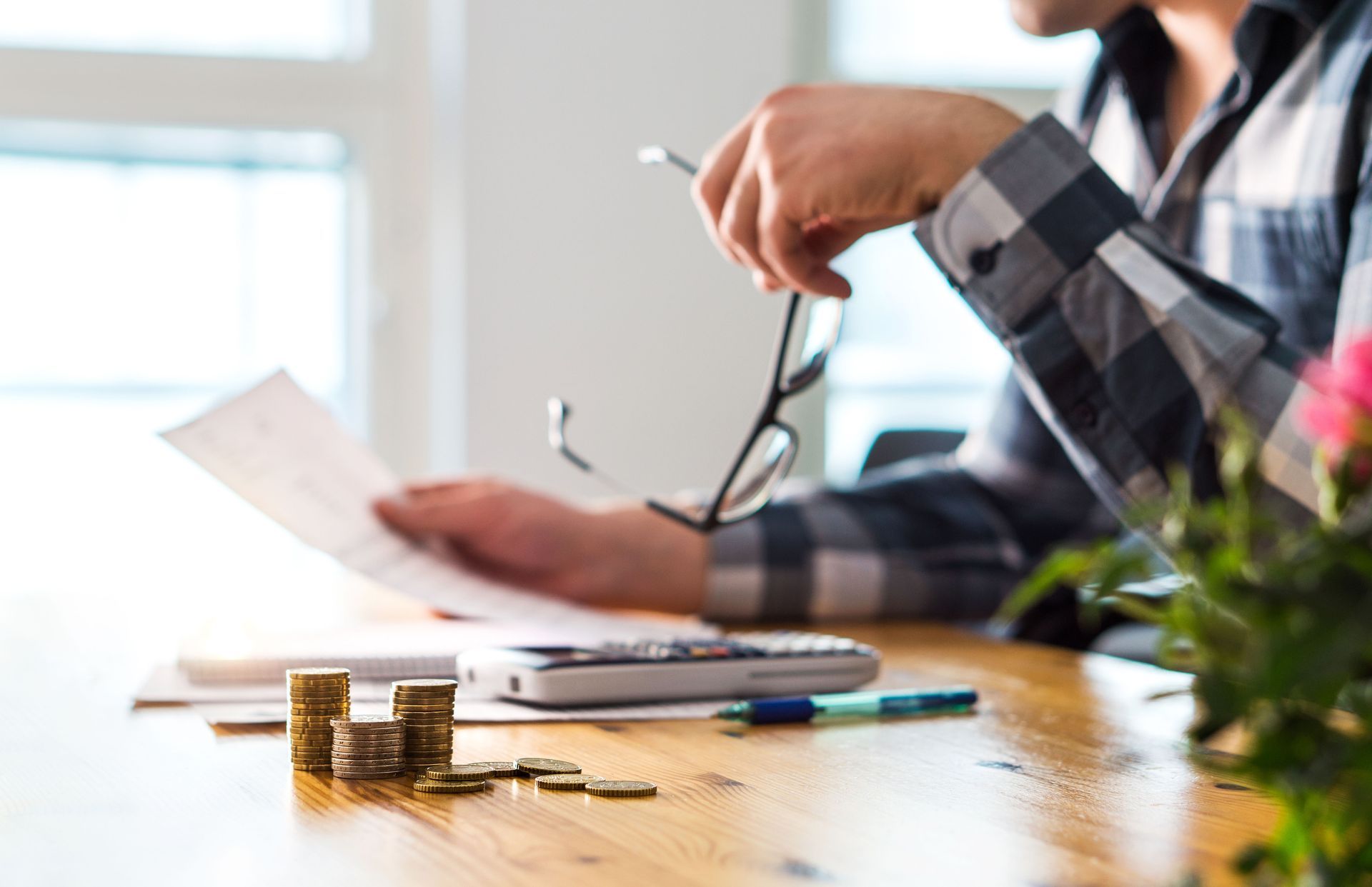 Person reviewing bills, holding glasses, with money, calculator, and pen on a wooden table.