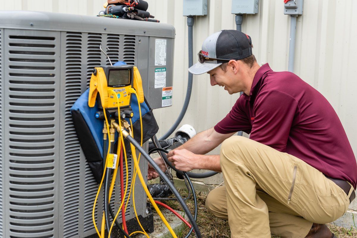 HVAC technician kneels, connecting gauges to an AC unit outside. He wears a maroon shirt, tan pants, and a baseball cap.