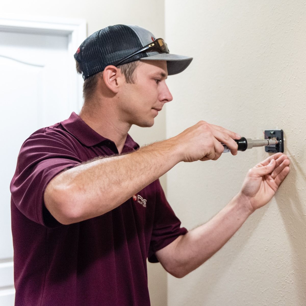 Man in a maroon shirt and hat installing something on a beige wall with a screwdriver.