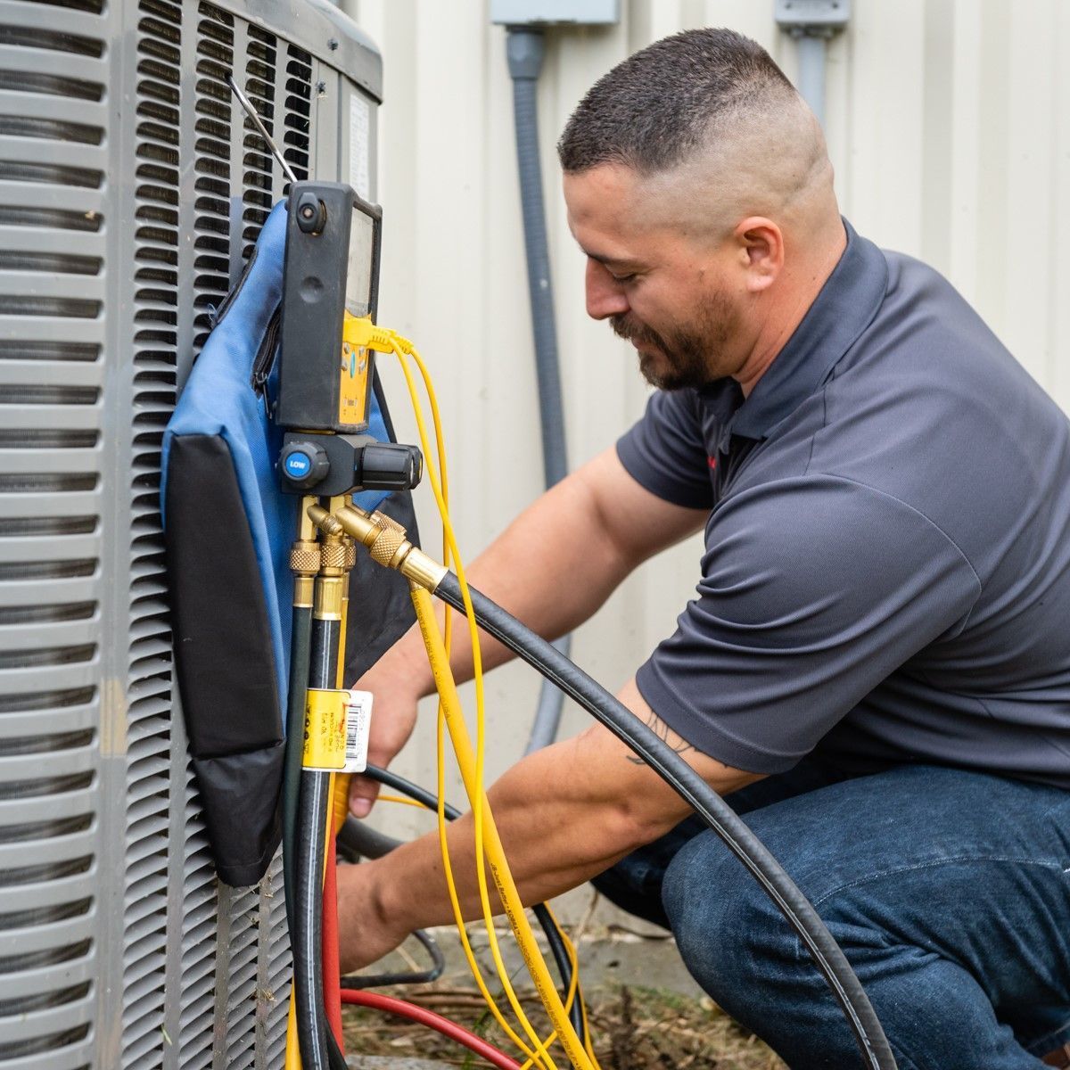 HVAC technician kneeling by outdoor unit, connecting hoses.  Wearing gray shirt and jeans, sunny outdoors.