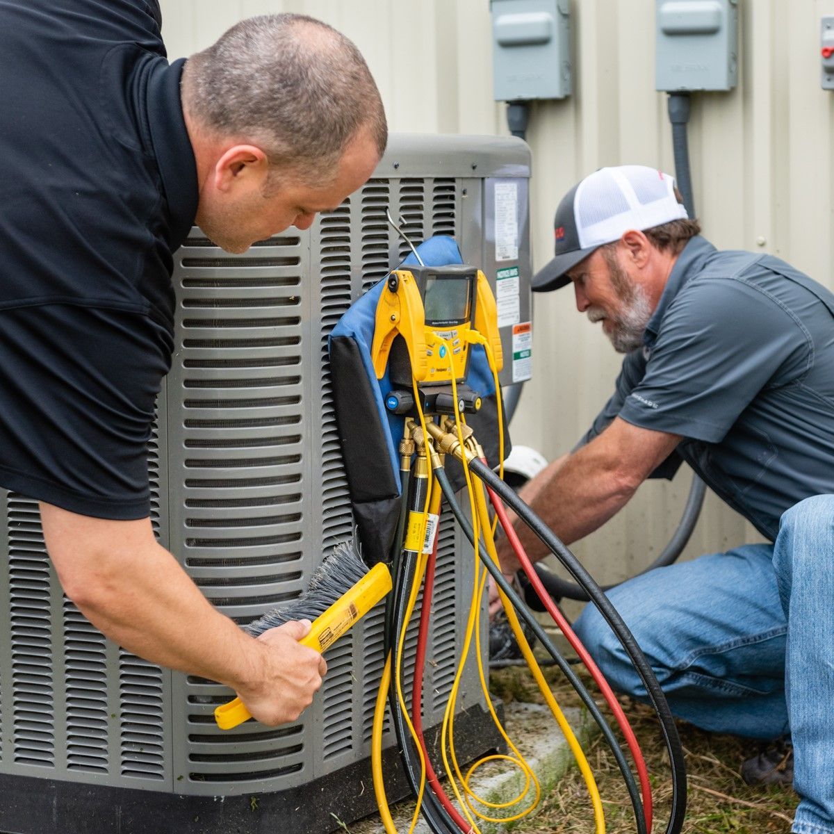 Two technicians working on an air conditioning unit outside, using tools and gauges.