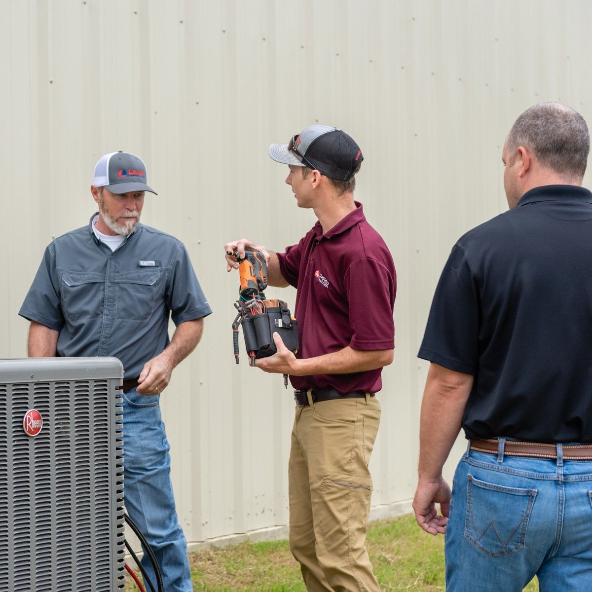 Three people inspecting an AC unit outdoors. One holds a tool, explaining to the other two.