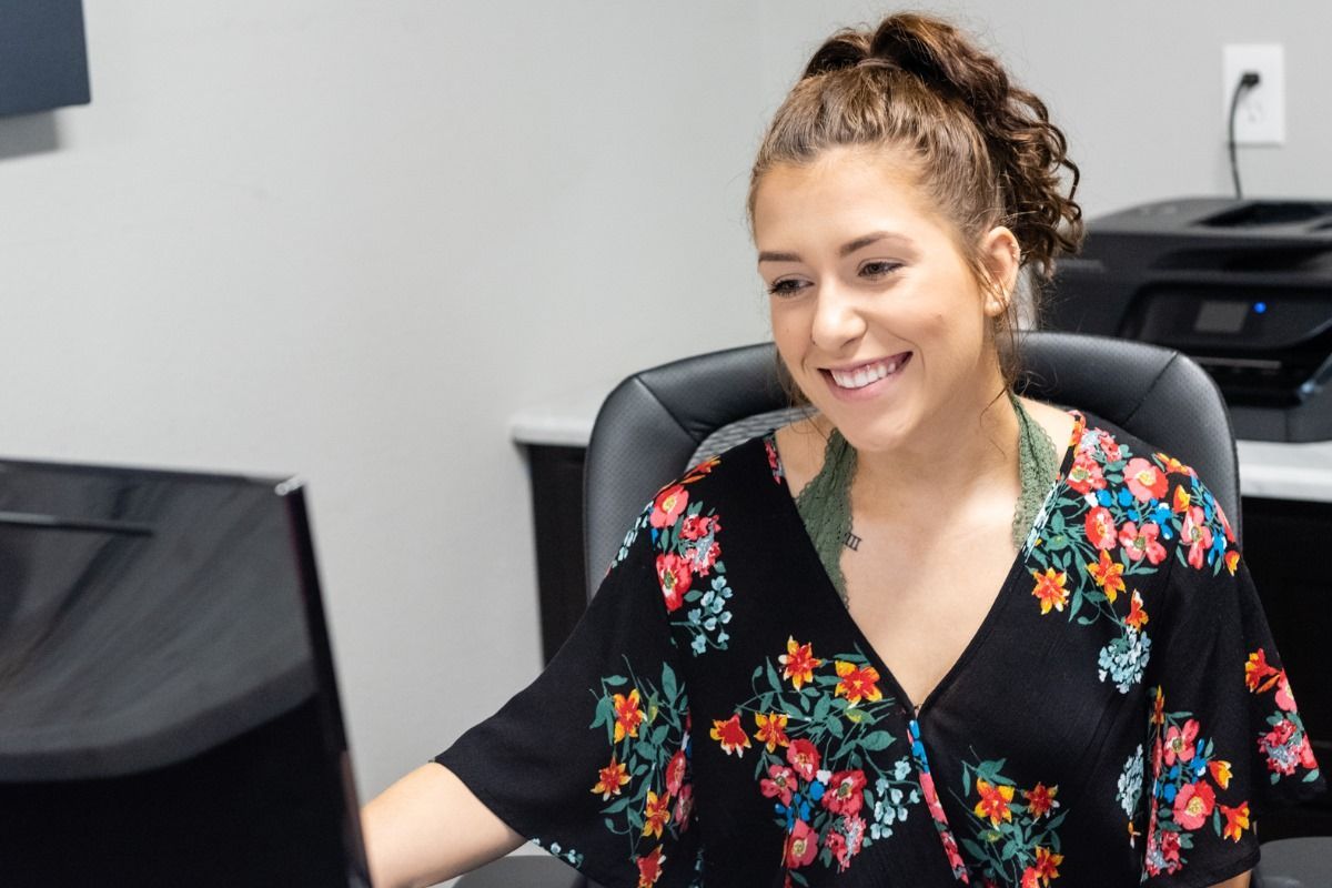 Woman with curly ponytail smiles, working at a computer in an office, wearing a floral top.