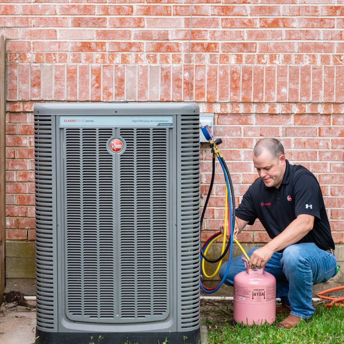 HVAC technician kneeling near a grey air conditioner, connecting hoses to a pink refrigerant tank. Brick wall backdrop.