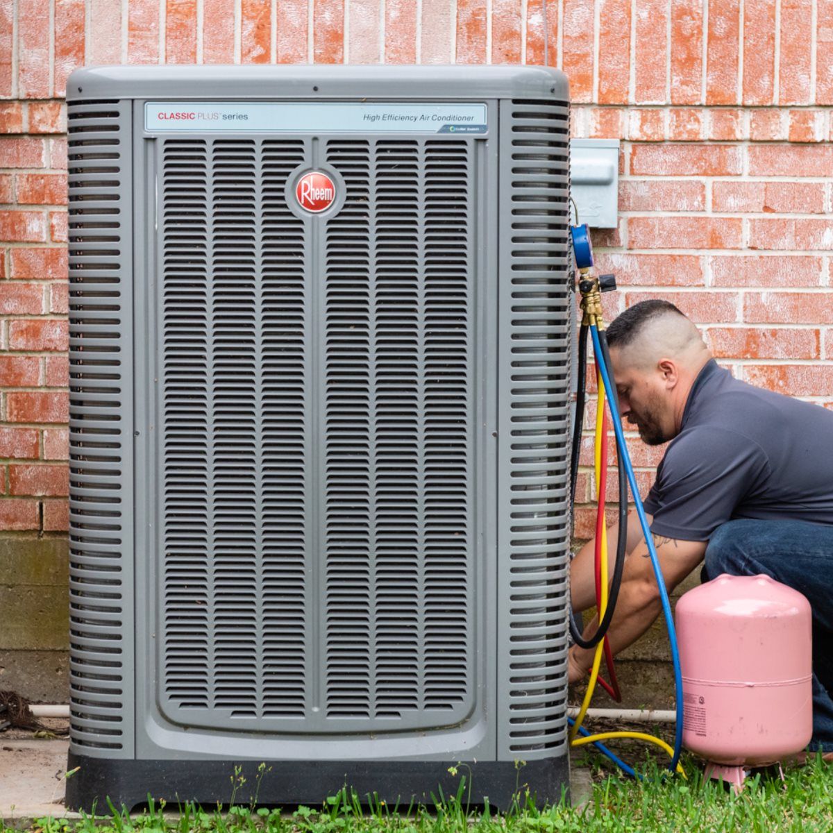 HVAC technician servicing a gray air conditioning unit against a brick wall, using gauges and a pink tank.