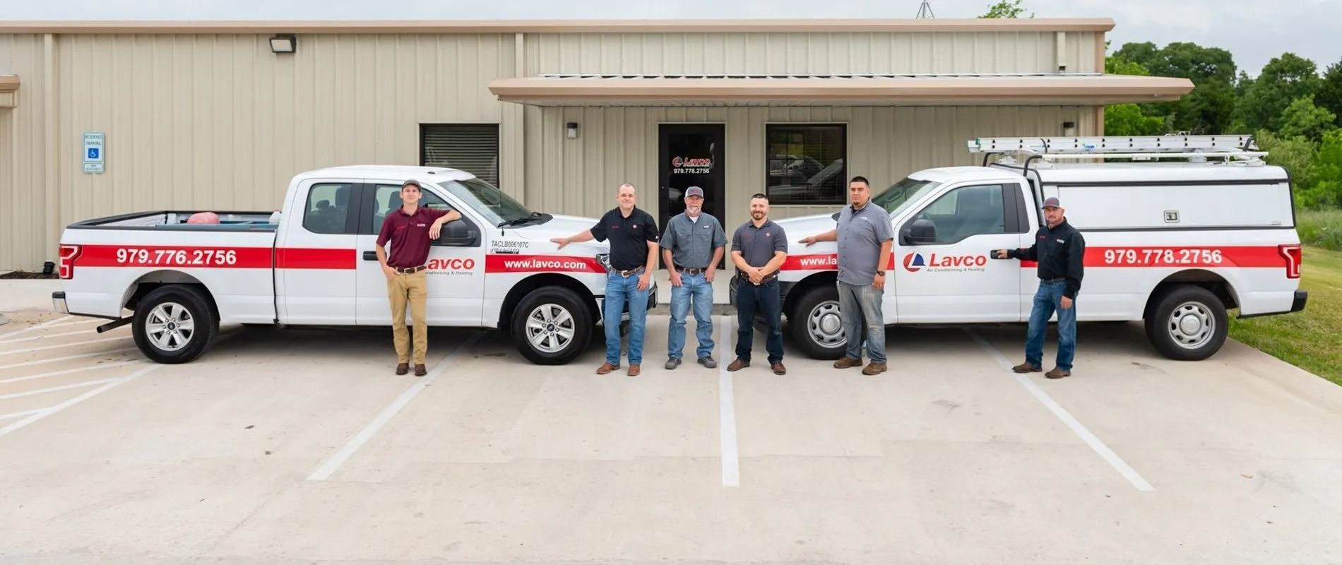 Six people stand in front of two white pickup trucks with red stripes, parked in front of a tan building.