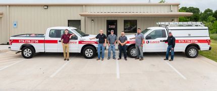 Six people stand in front of two white pickup trucks with red stripes, parked in front of a tan building.
