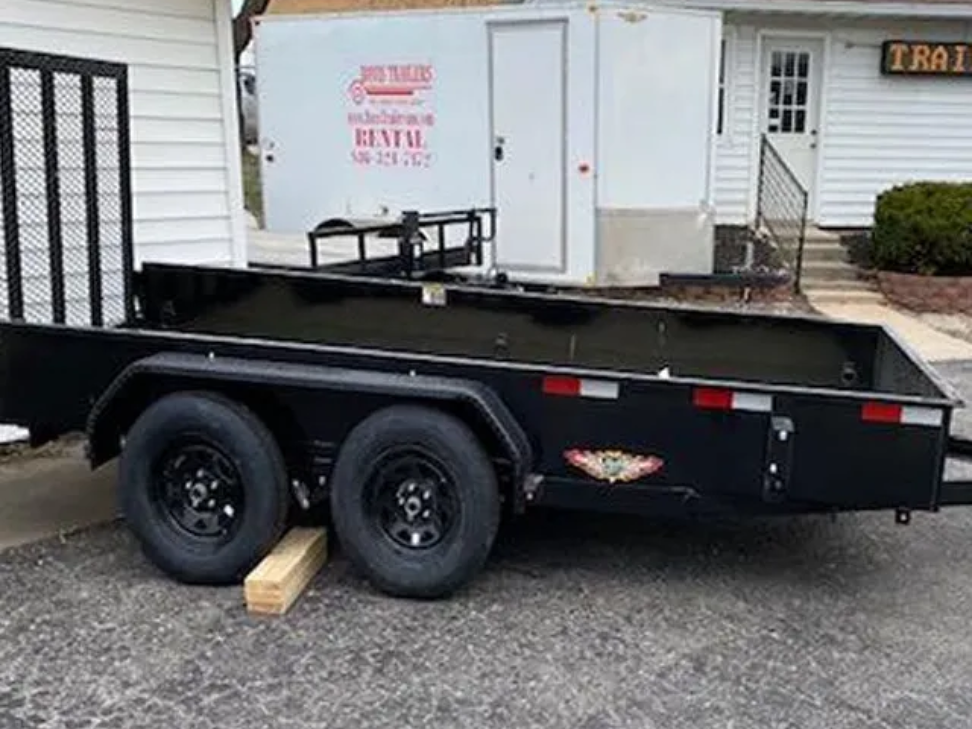 Black utility trailer parked in front of a white building, with two wheels and a wooden block.