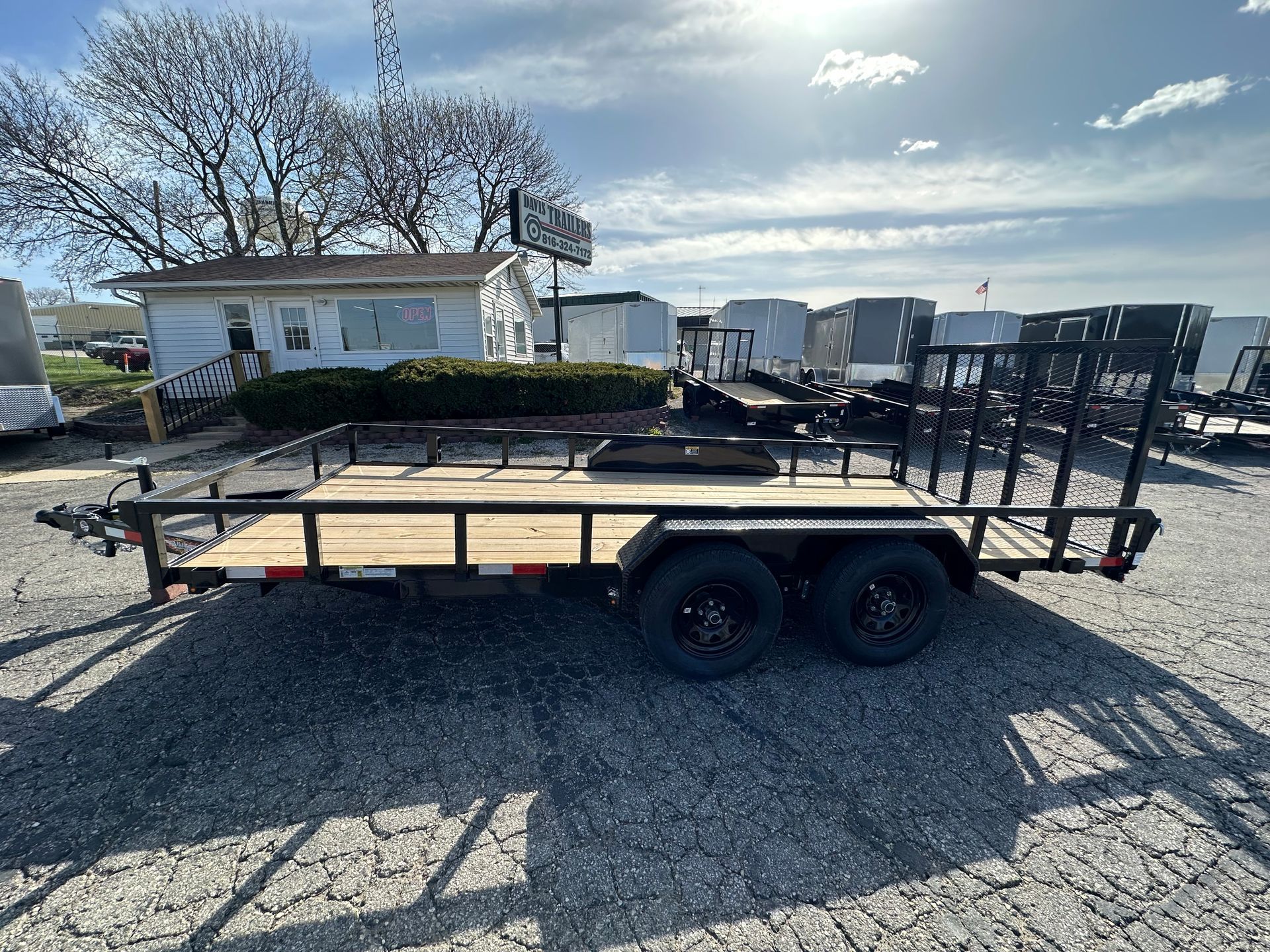 Black utility trailer with dual wheels, parked on asphalt near a building and other trailers. Sunny day.