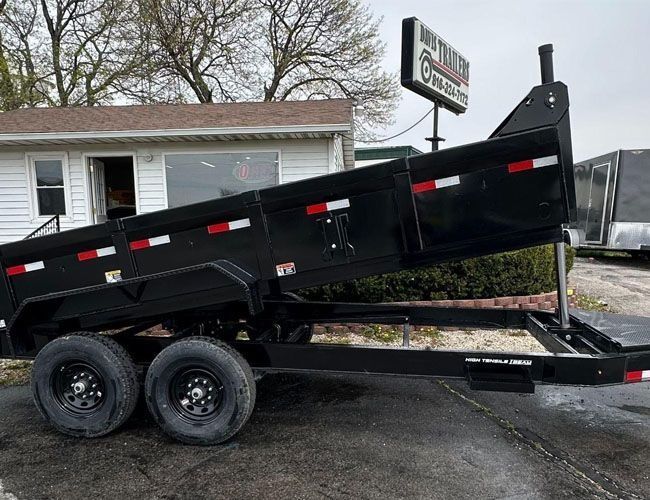 Black dump trailer with raised bed, two wheels, and a business sign in the background.