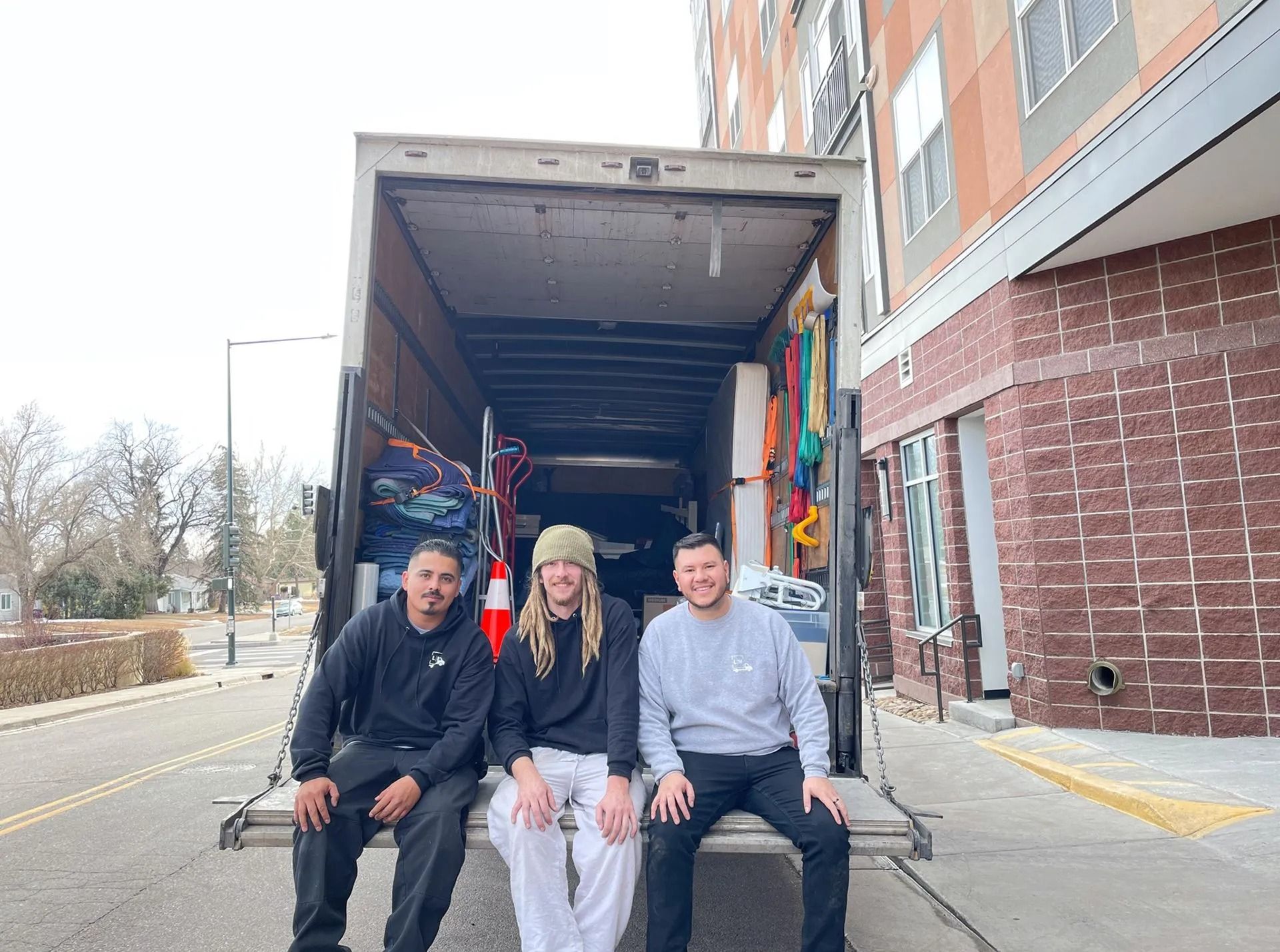 Three people are sitting on the back of a moving truck.