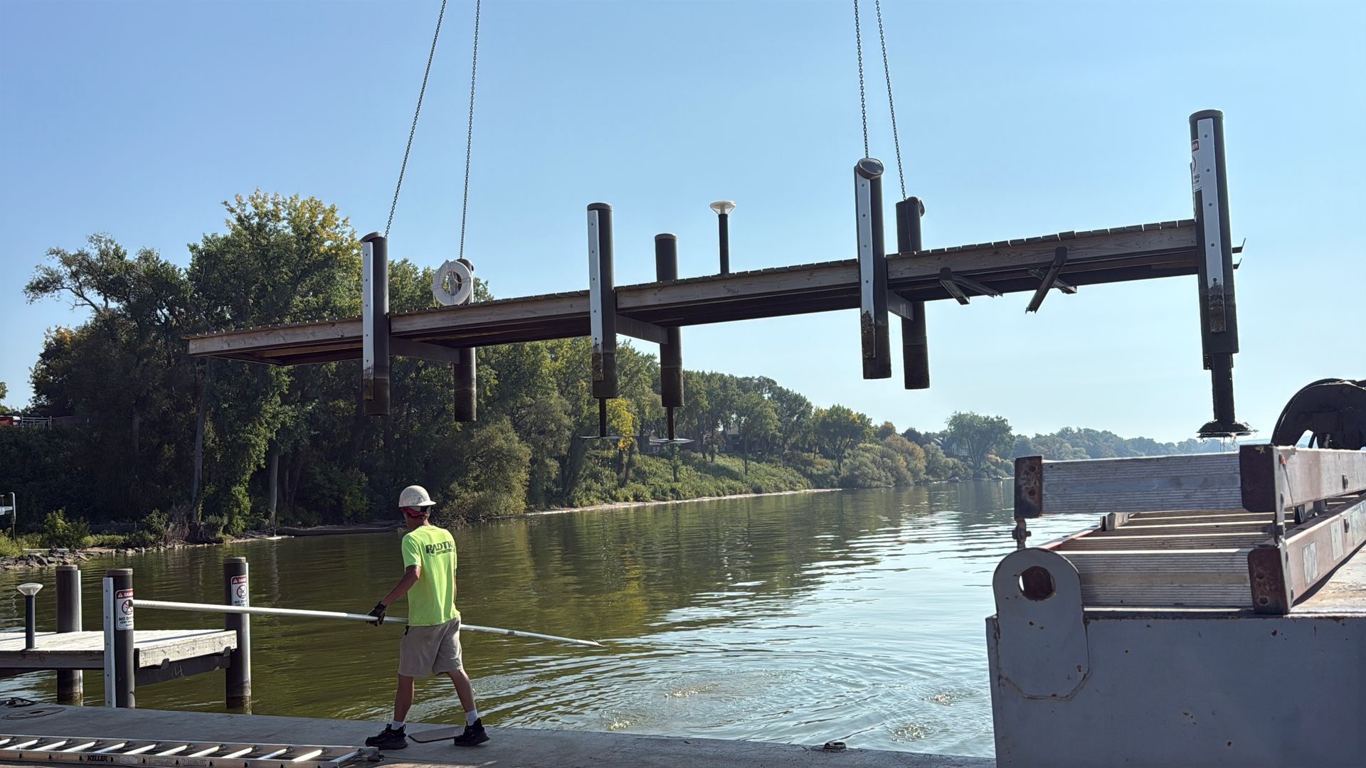 Man in safety vest guiding the removal of a dock section with a crane over a river.