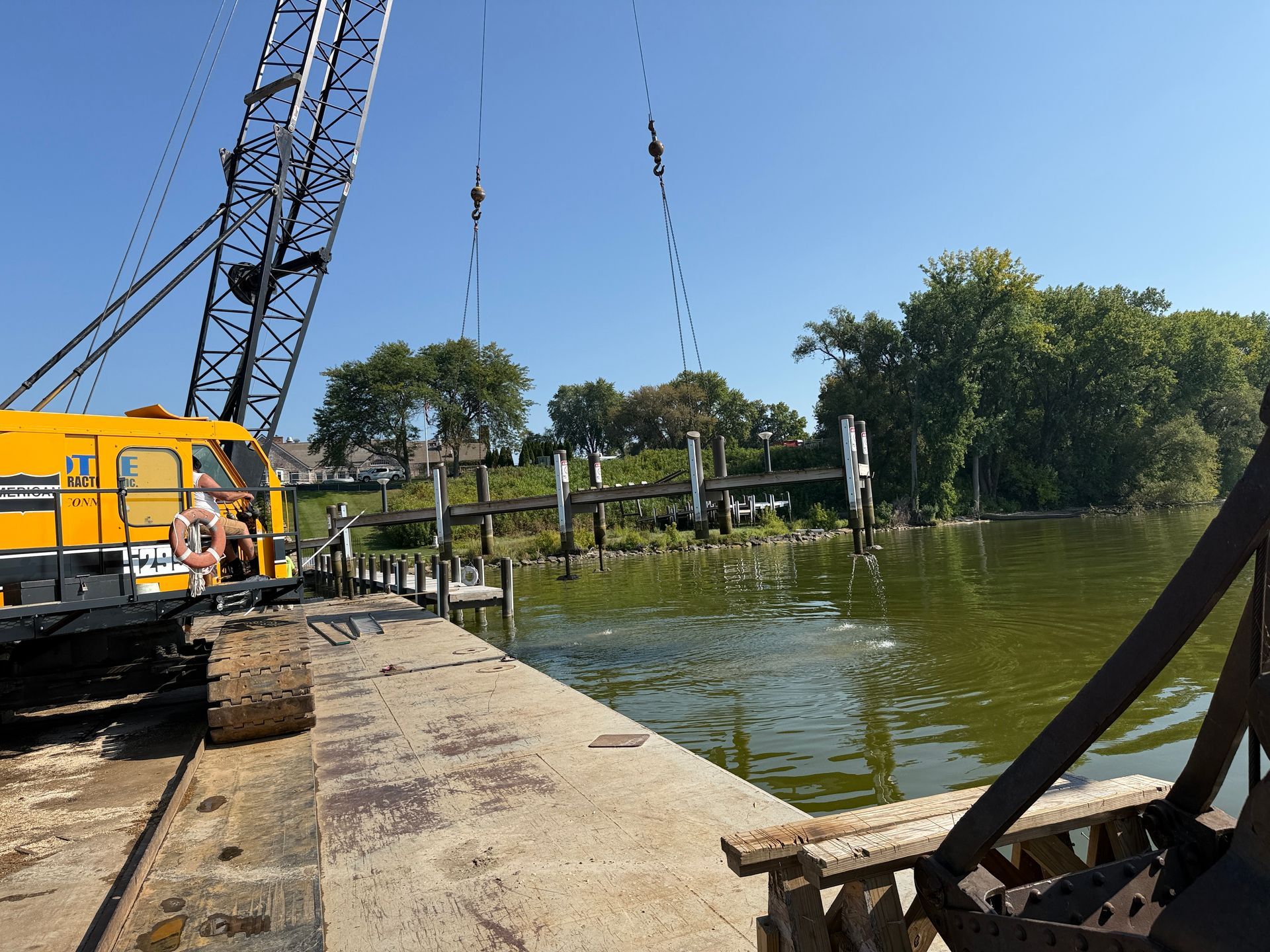 A crane is lifting debris from a murky river. Dock pilings and trees line the opposite bank.