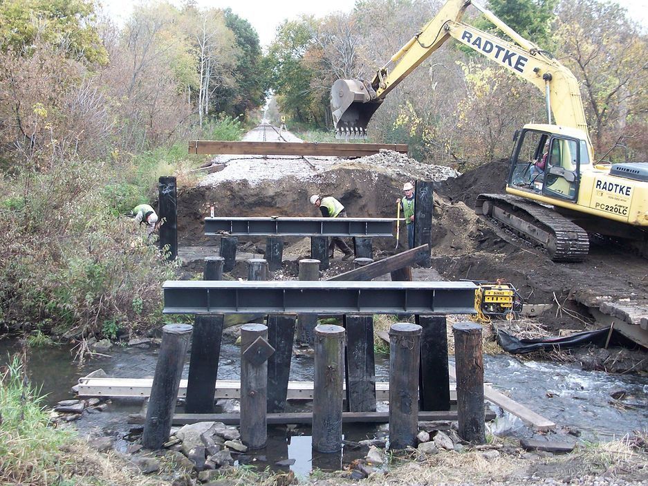 A yellow excavator is working on a bridge over a river.