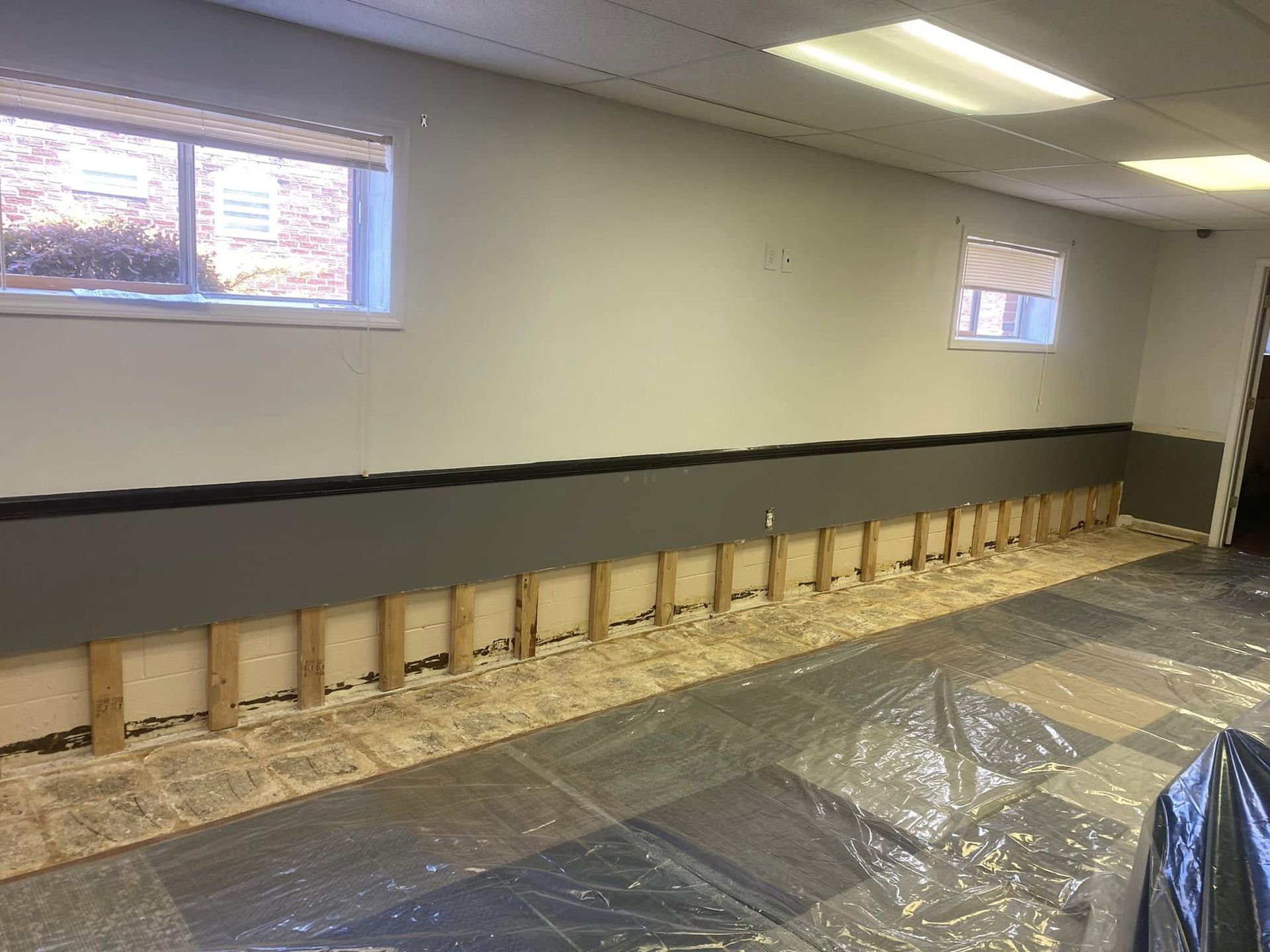 Room with exposed wall studs, gray and white walls, two windows, and plastic sheeting on the floor.