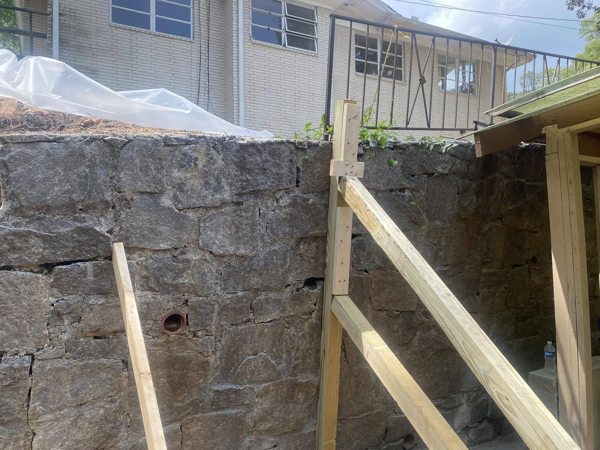 Stone wall with wooden support structure in front; house in background.