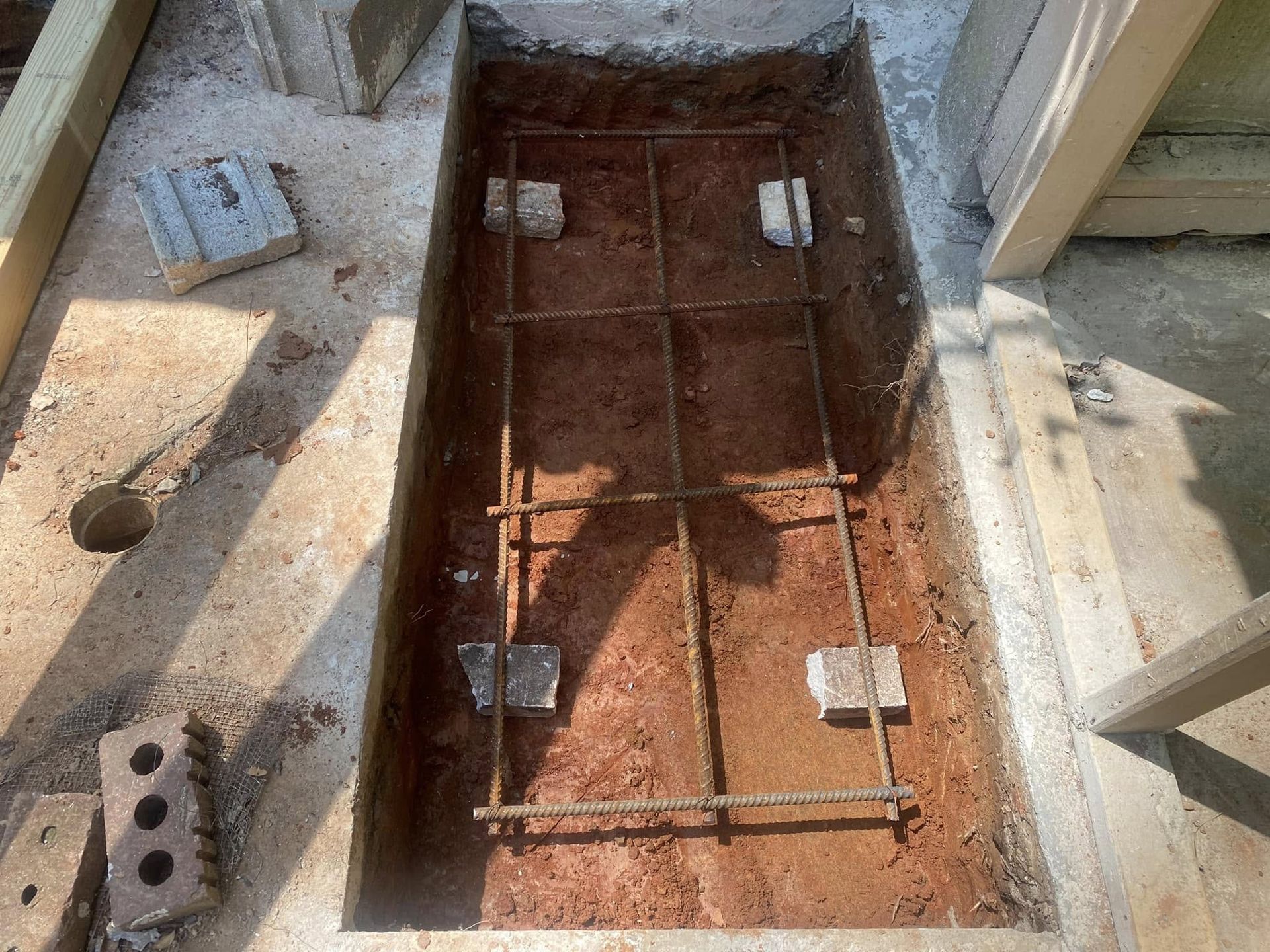 Excavated rectangular pit with steel reinforcing bar grid and concrete blocks for footings.