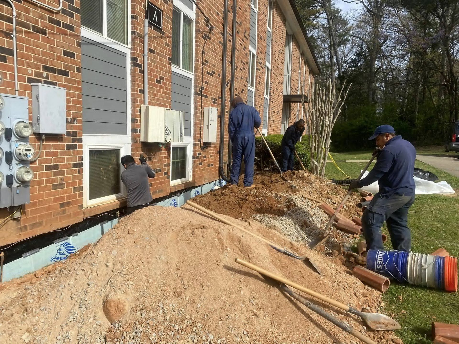 Construction workers digging near a brick building's foundation. Piles of dirt and equipment are visible.