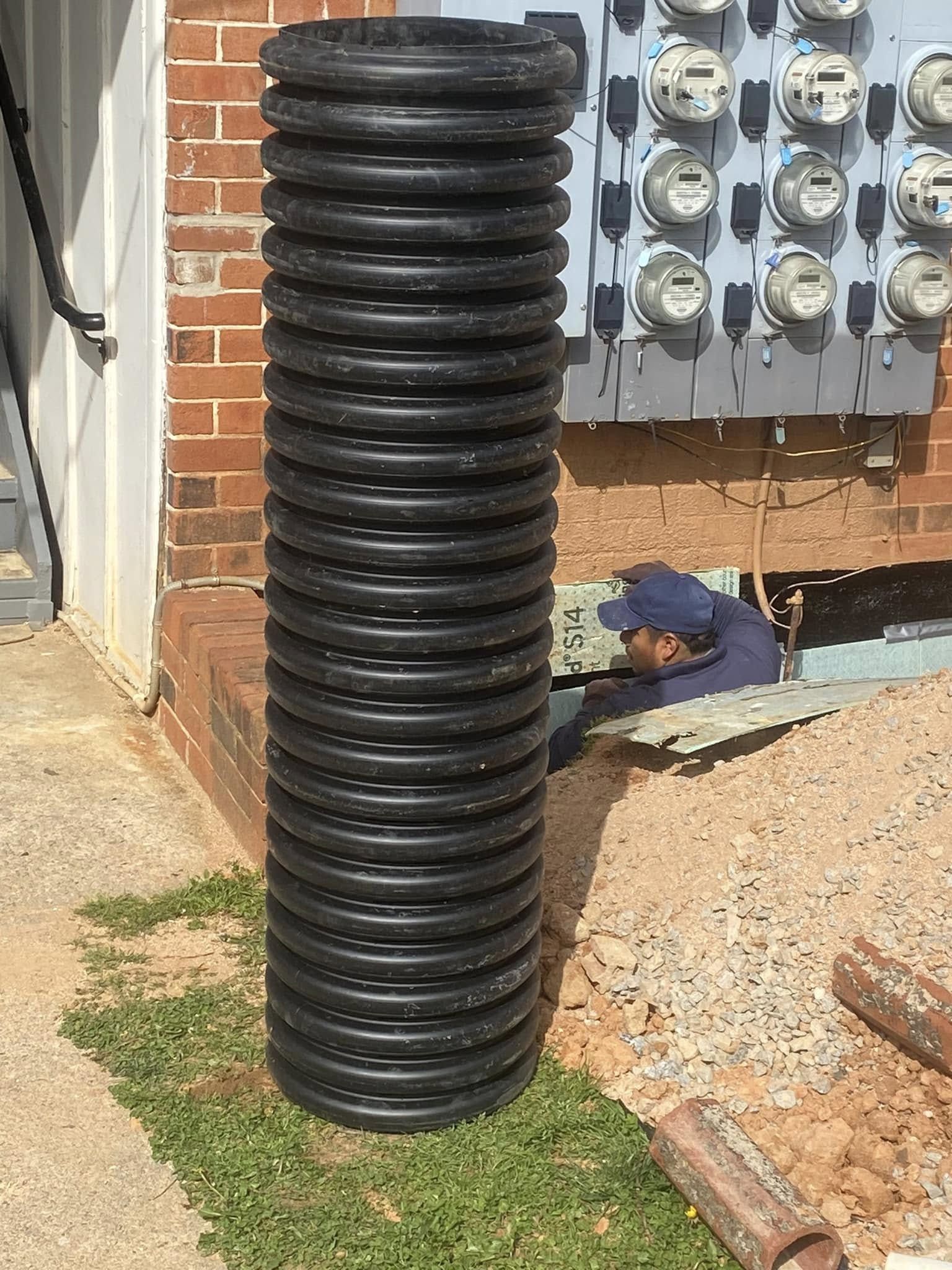 Black corrugated pipe standing upright next to a brick building. A person is visible in a crawlspace.