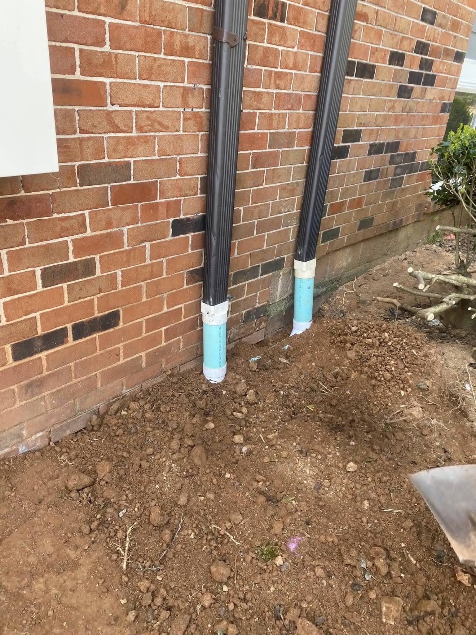 Two black downspouts attached to a brick wall, leading into blue PVC pipes in the dirt.