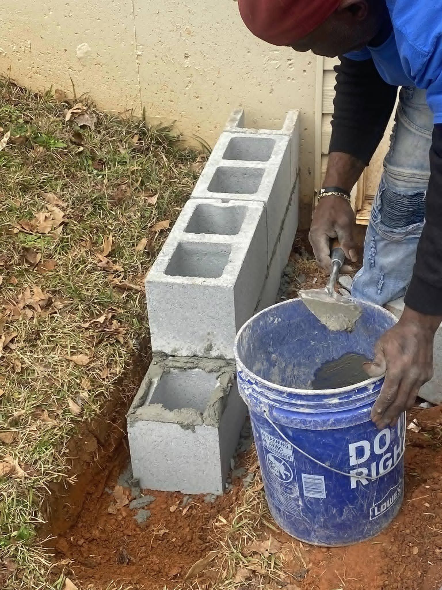 Man laying cinder blocks, using a trowel and bucket of cement, beside a wall and grassy area.