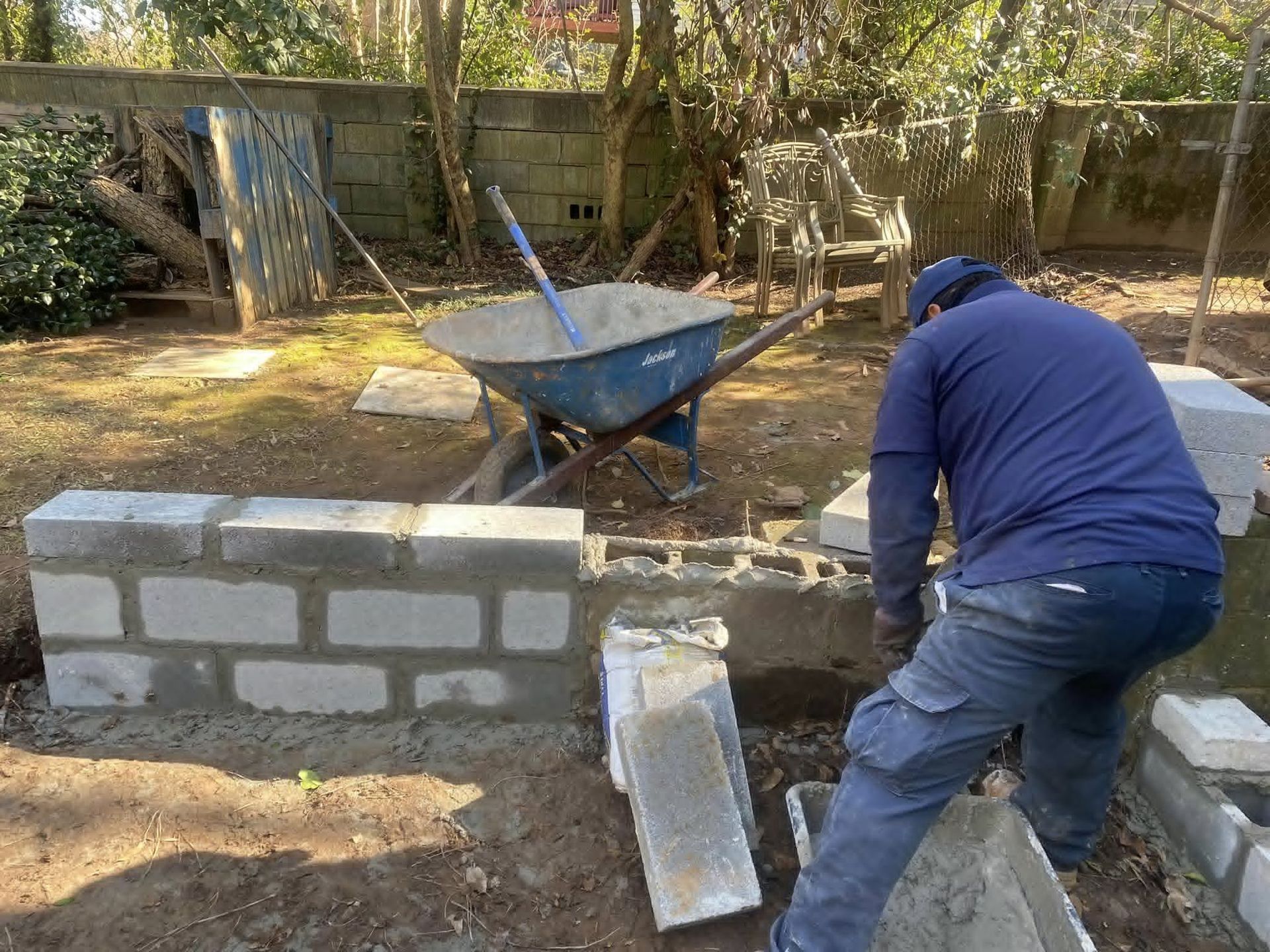 Person building a concrete block wall in a backyard with a wheelbarrow.