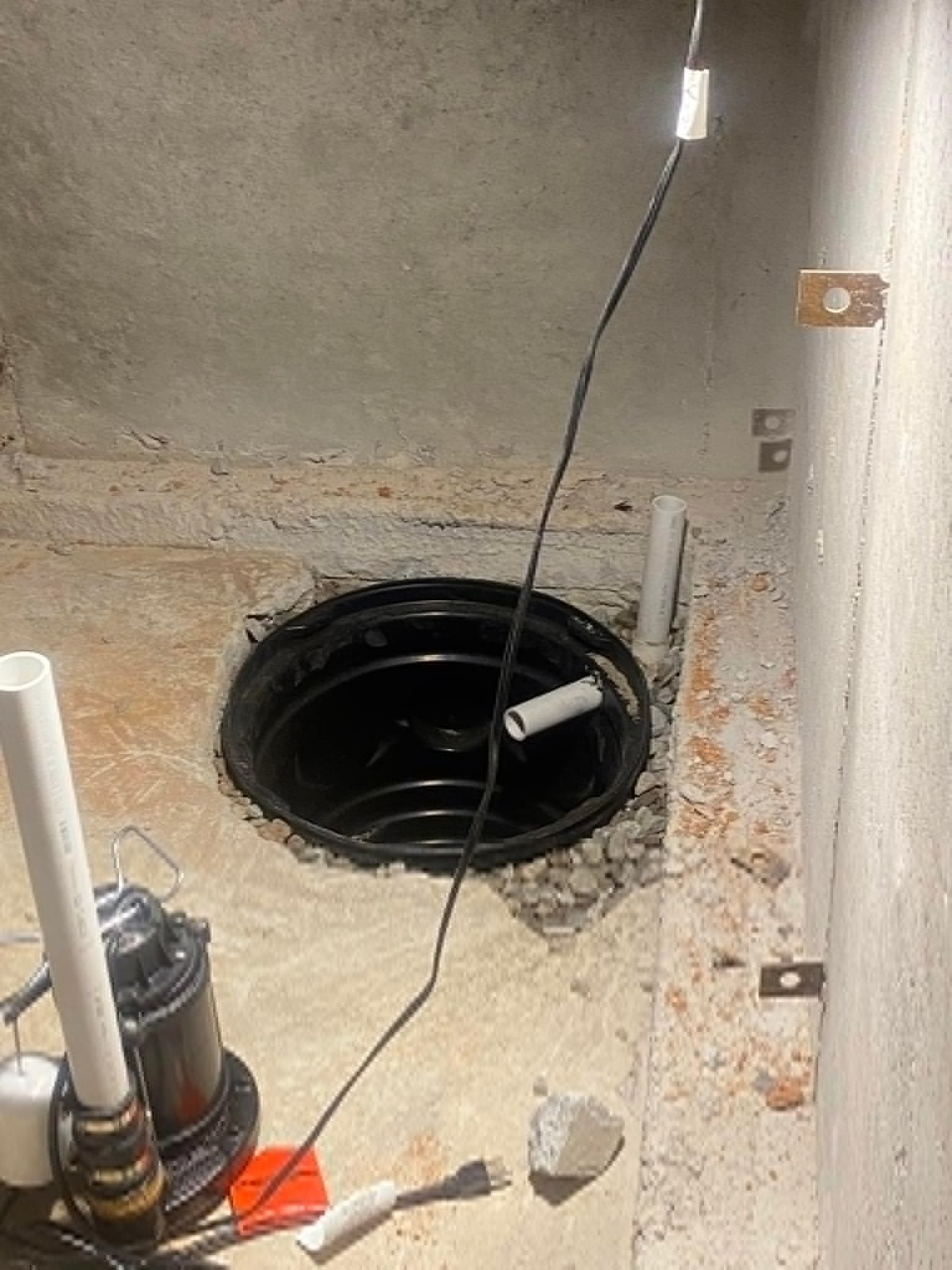 Sump pump in a concrete pit. Black pump, white pipes, gray concrete walls. Electrical cord present.