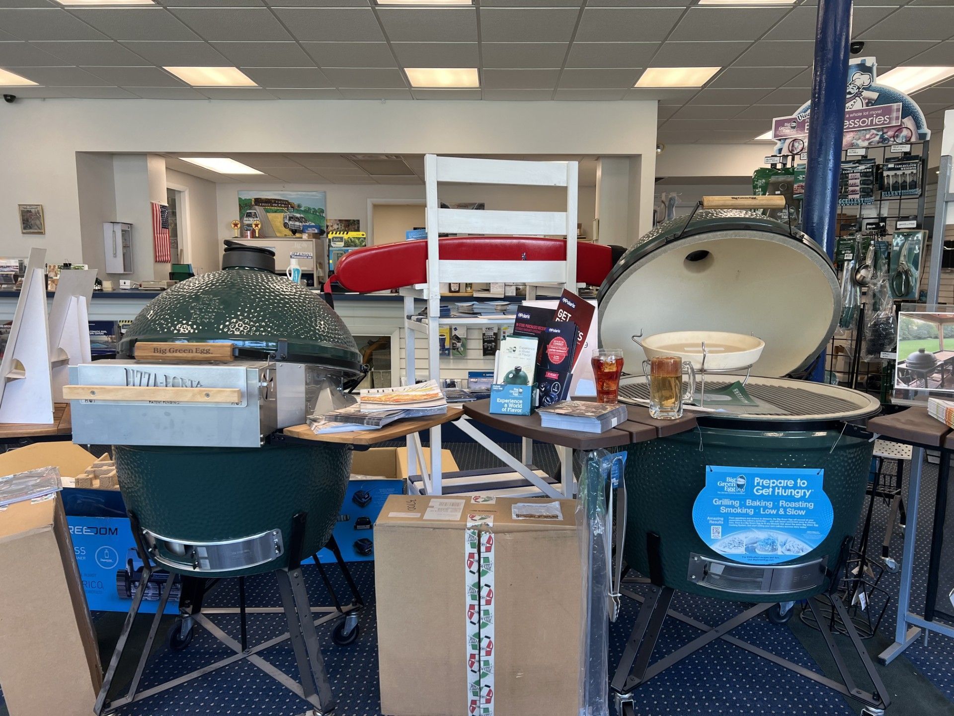 Indoor store display of two large green grills with open lids and assorted outdoor supplies on tables