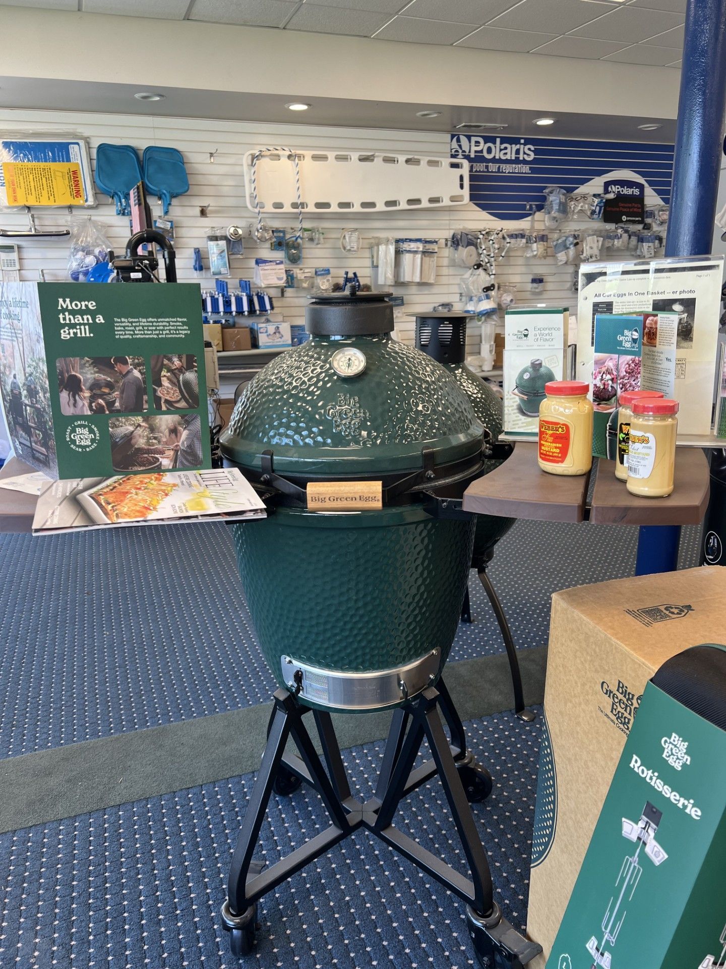 Green kamado grill on display at a store, with a table of food and drinks behind it.