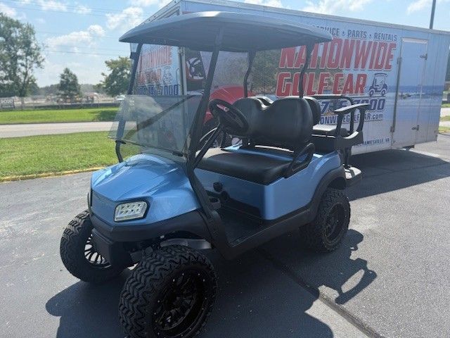 Blue golf cart with black wheels parked on pavement, in front of a trailer with