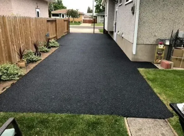 Black asphalt driveway next to a light-colored house and a wooden fence. Green grass borders the driveway.