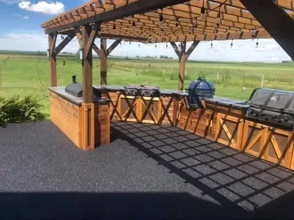 Outdoor kitchen with a wooden pergola, multiple grills, and cabinets, set against a green field.
