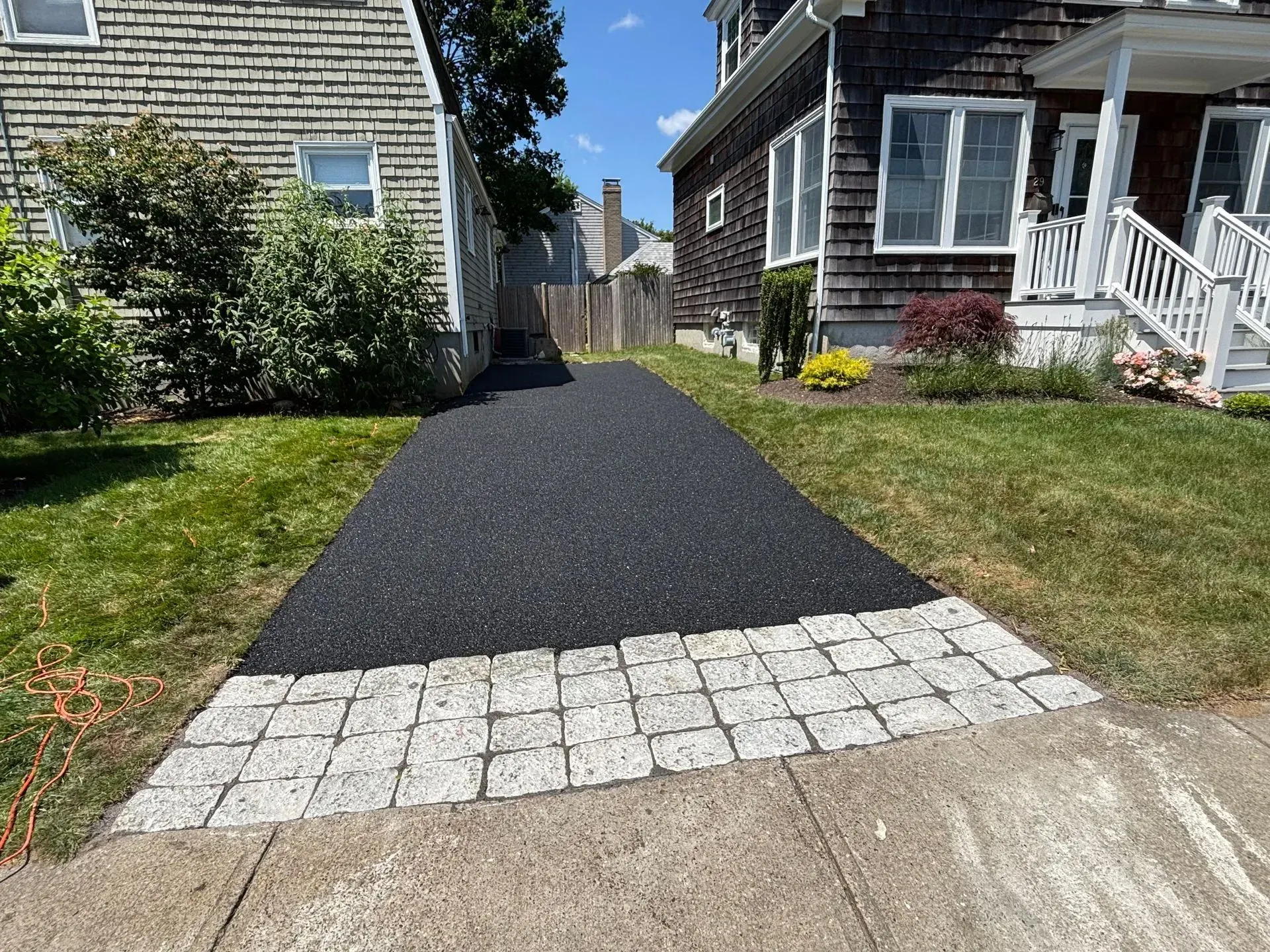 Black asphalt driveway with light-colored brick border, between two houses.