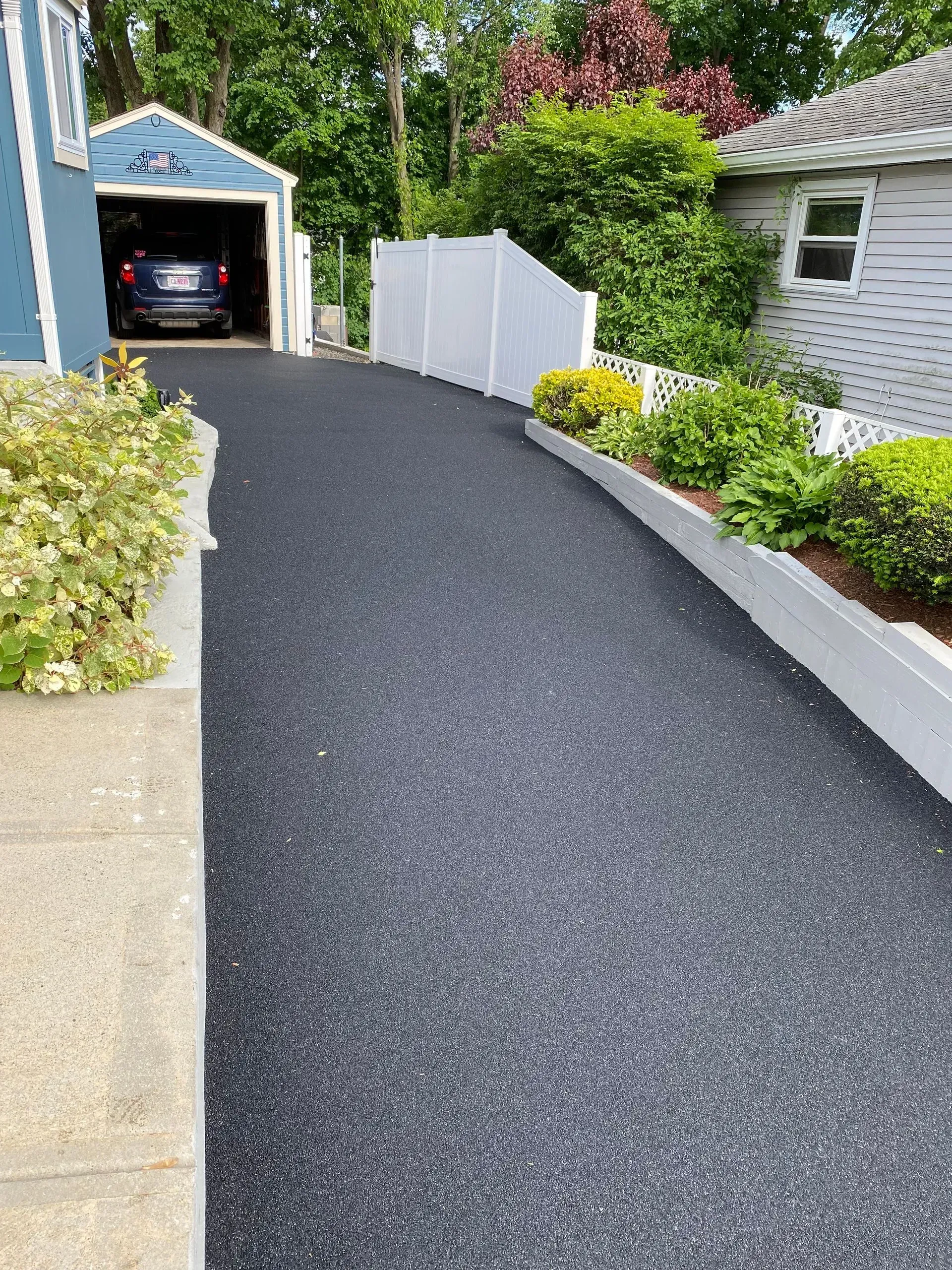 Asphalt driveway leading to a blue garage; white fence and garden beds border the drive.