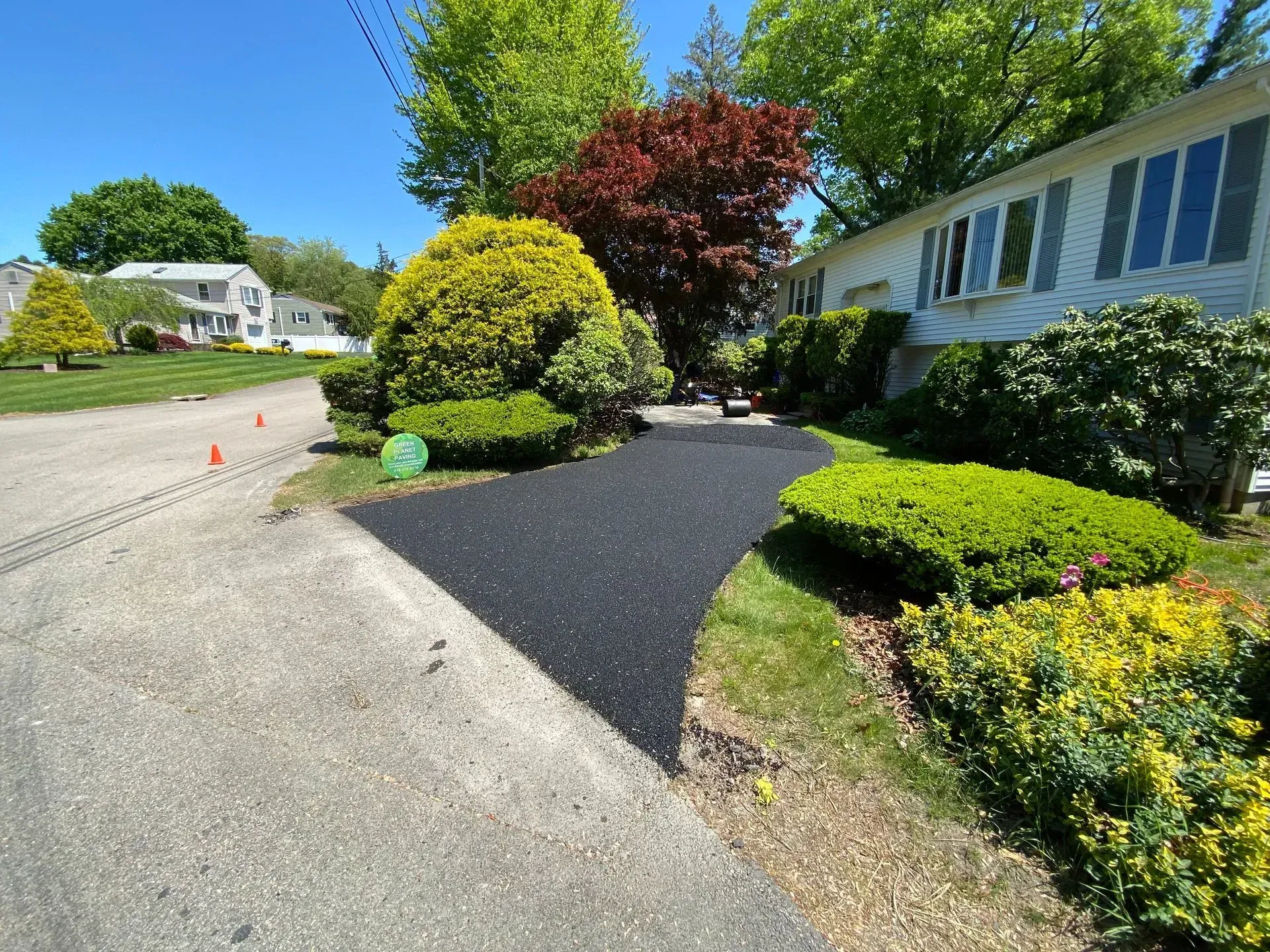Black asphalt driveway curves to a house with white siding and lush landscaping on a sunny day.