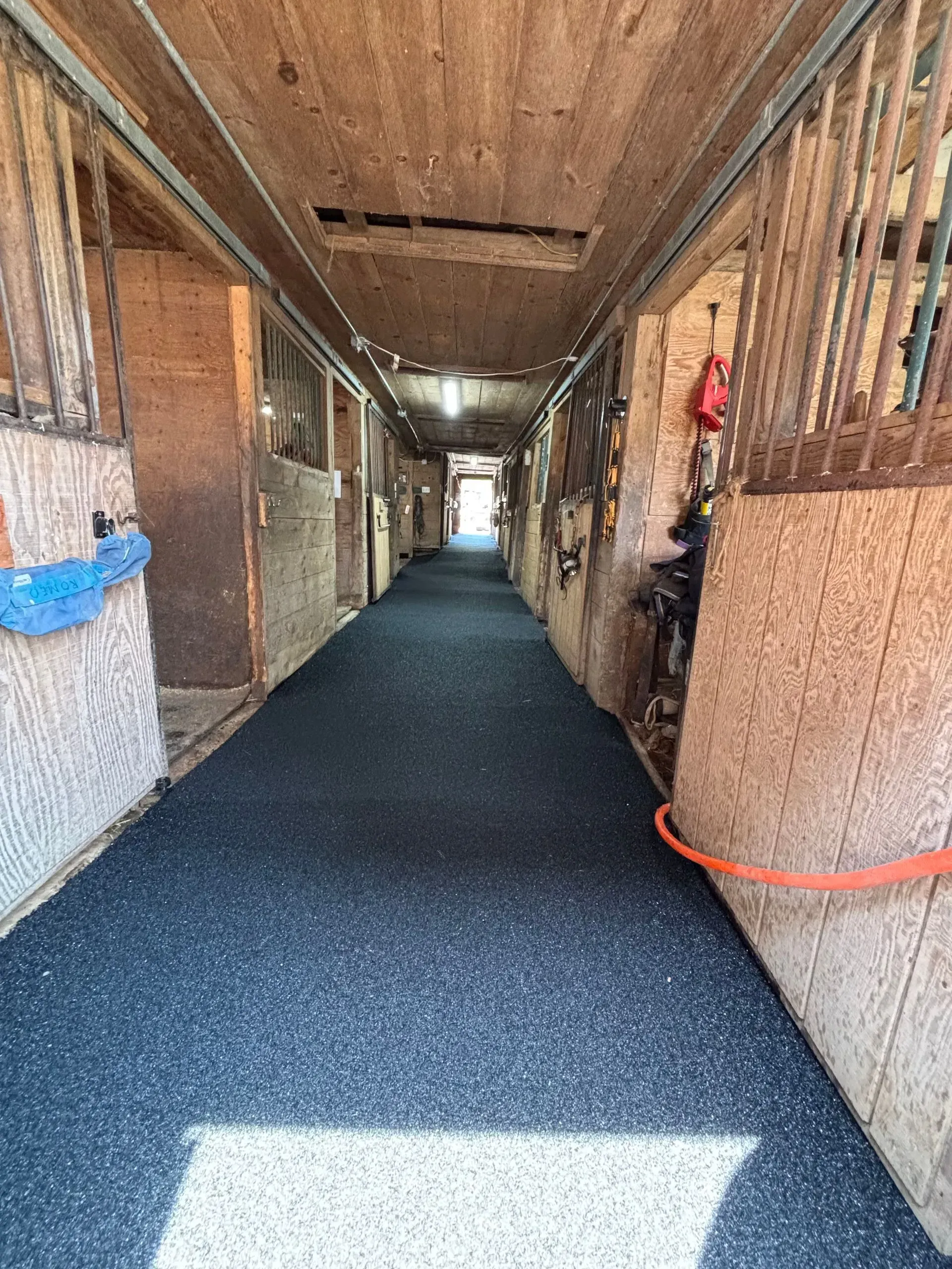 Long hallway in a stable with stalls on either side and black floor covering. Wooden walls and roof.