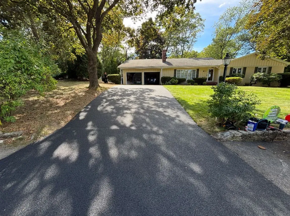 Asphalt driveway leading to a one-story beige house with a two-car garage, surrounded by trees and grass, on a sunny day.
