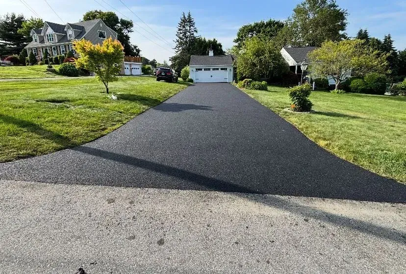 Paved driveway leading to a garage, flanked by green lawns and houses on a sunny day.