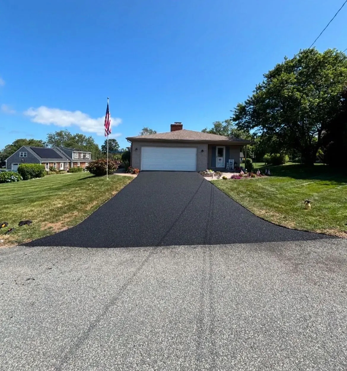 Newly paved asphalt driveway leading up to a beige house under a blue sky, an American flag visible.