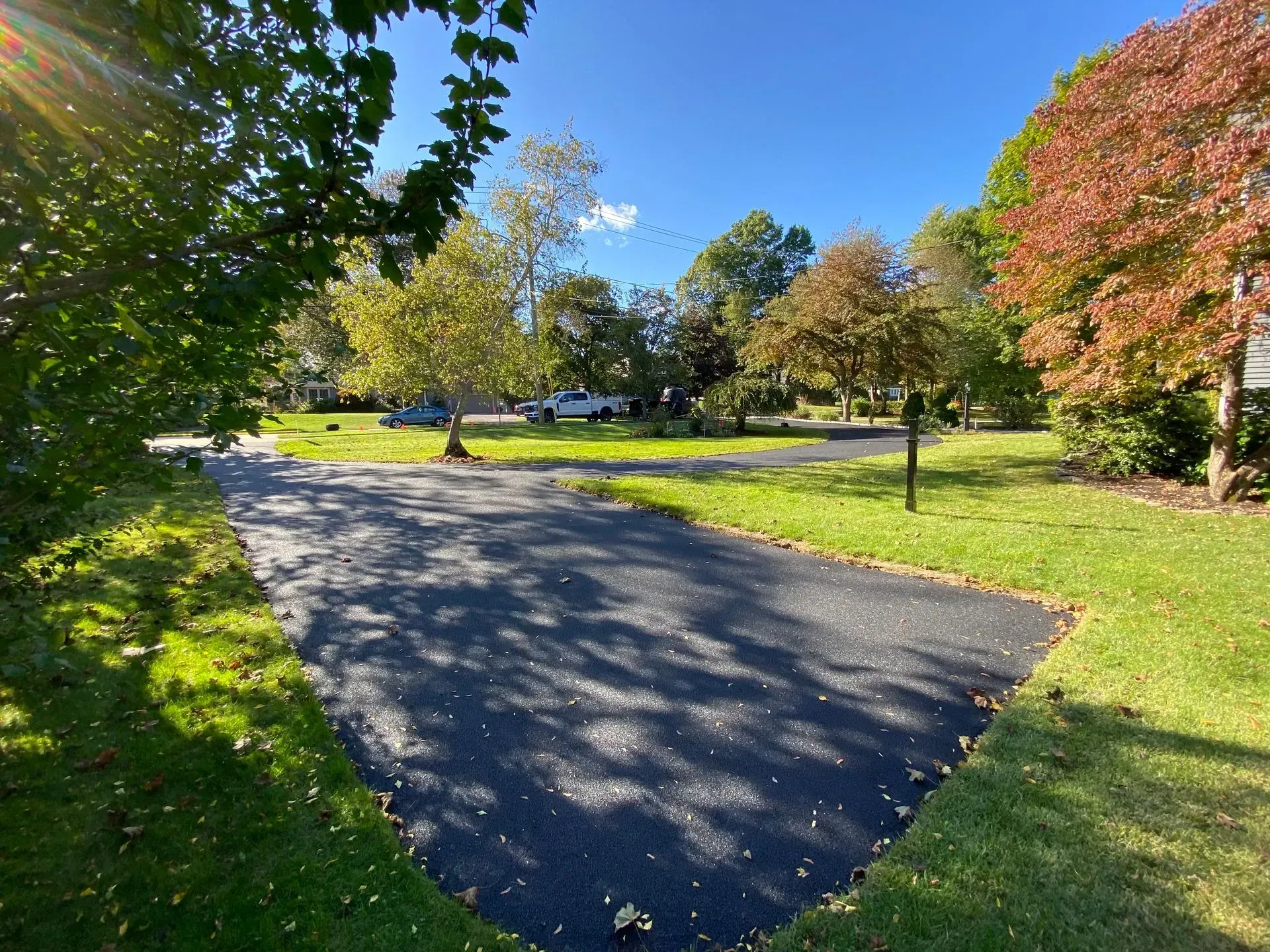 Asphalt driveway surrounded by green grass and trees with fall foliage under a clear blue sky.