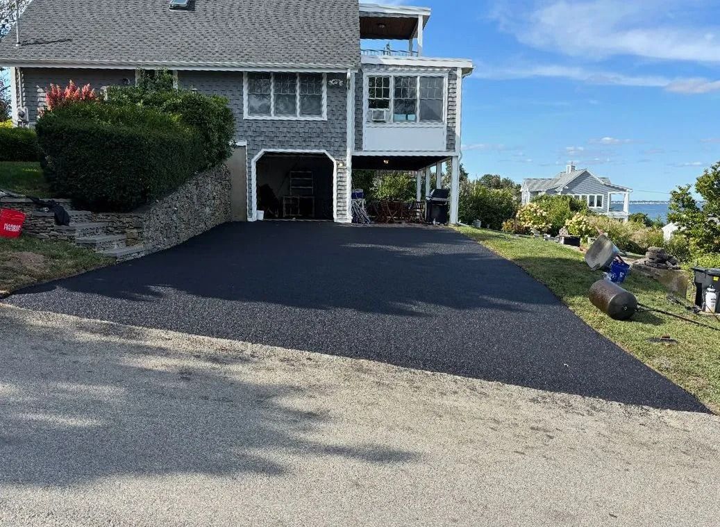 Asphalt driveway in front of a gray house, partially surrounded by grass and a stone wall.