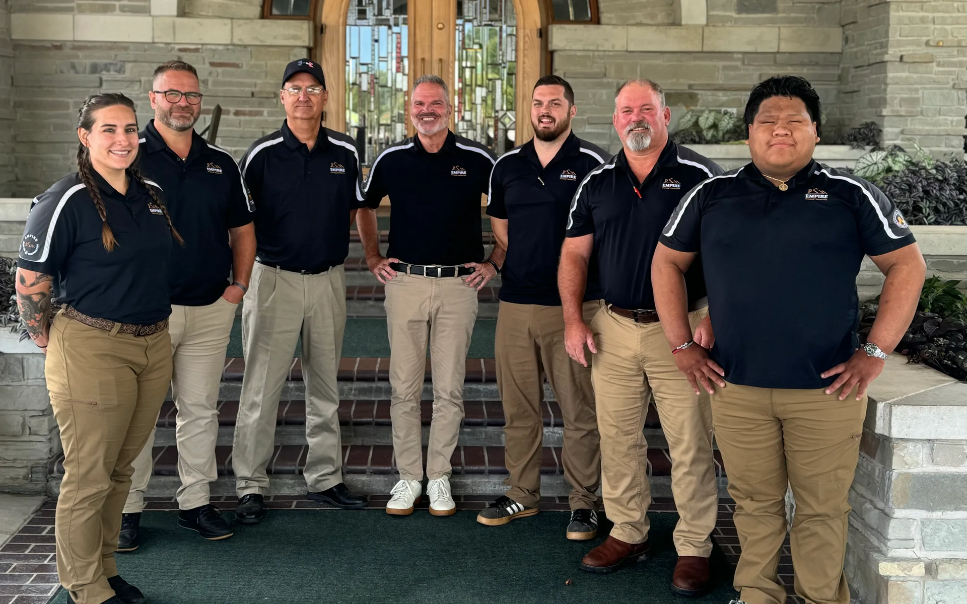 Seven people in black shirts and khaki pants stand outside a building, smiling.