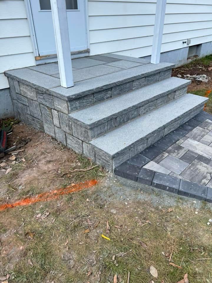 Stone steps with white railings leading to a gray house porch, beside a paved walkway and dirt yard.