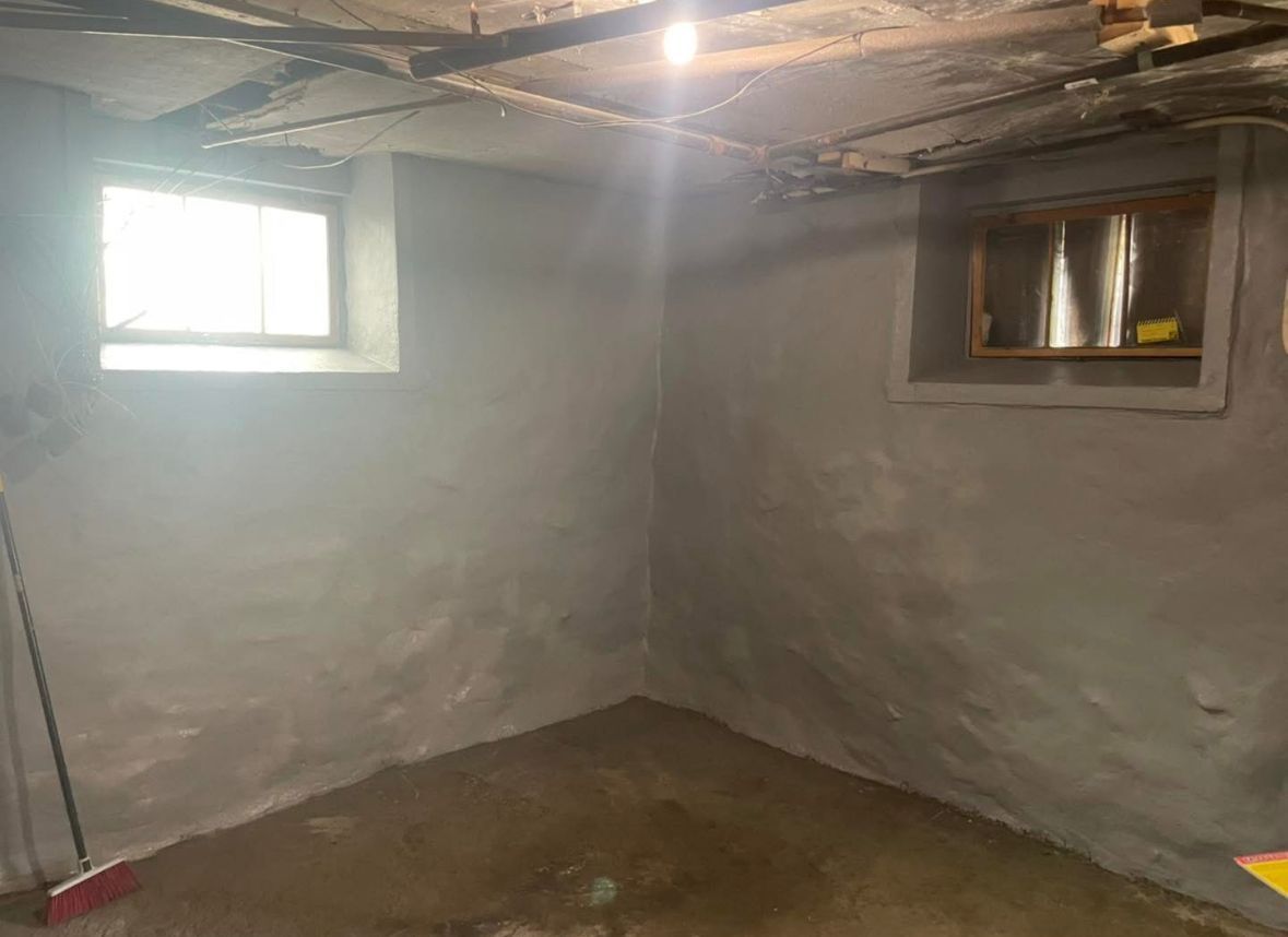 Bare basement room with gray walls, exposed ceiling beams, two small windows, and a concrete floor.