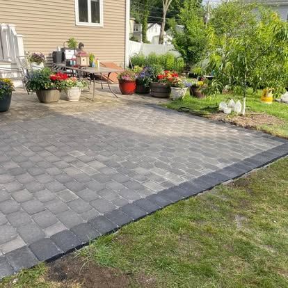Paved patio beside a house with potted flowers and garden beds in a sunny yard.
